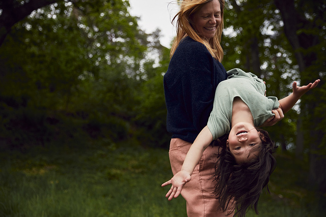 Woman playfully holding a smiling child upside down in a green forest setting.