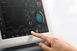A hand adjusting settings on a medical monitor displaying graphs, numbers, and a circular gauge.