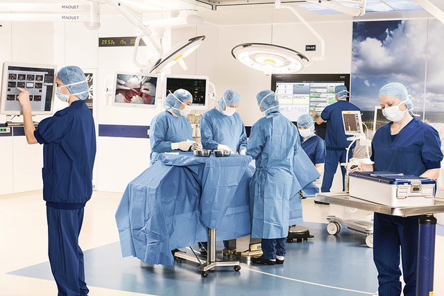 Medical team in blue scrubs performing surgery in a bright operating room with monitors and medical equipment.